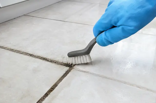 Stiff brush scrubbing dark grout lines between white ceramic floor tiles